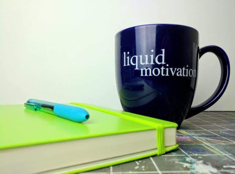 Motivation coffee mug with "liquid motivation" text and a lime green notebook with pen on table.